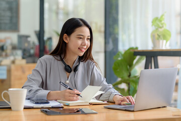 Beautiful Asian businesswoman taking notes on her laptop computer having fun and smiling happily at work.