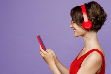 Side view young smiling woman 20s she wear red tank shirt eyeglasses headphones listen to music hold in hand use mobile cell phone isolated on plain purple backround studio. People lifestyle concept.