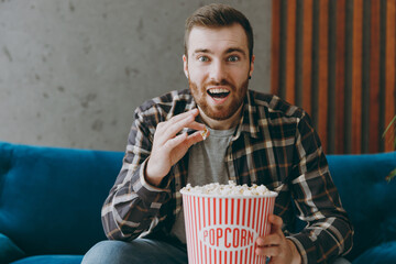 Young shocked excited man 20s he wears brown shirt hold takeaway bucket on popcorn watch tv show comedy movie film sitting on blue sofa in own living room apartment stay home indoor flat on weekends.