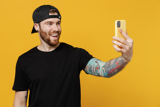 Young smiling bearded tattooed man 20s he wears casual black t-shirt cap doing selfie shot on mobile cell phone post photo on social network isolated on plain yellow wall background studio portrait