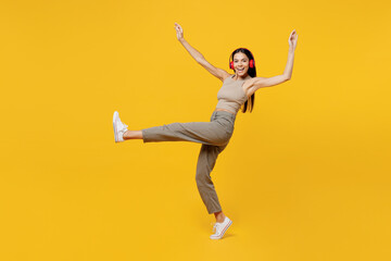Full body happy young latin woman 30s she wear basic beige tank shirt headphones listen to music dance raise up hands leg isolated on plain yellow backround studio portrait. People lifestyle concept.