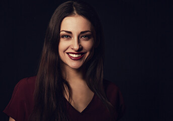 Beautiful young business toothy smiling woman thinking and looking happy in burgundy blouse and with red lipstick on dark shadow black background with empty copy space. Portrait closeup front center