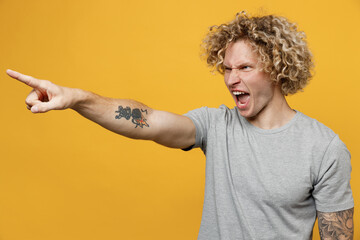 Young indignant furious sad dissatisfied displeased caucasian man 20s he wear grey t-shirt point index finger aside scream isolated on plain yellow backround studio portrait. People lifestyle concept.