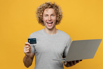 Young happy caucasian man 20s he wear grey t-shirt using laptop pc computer hold credit bank card doing online shopping order delivery booking tour isolated on plain yellow backround studio portrait