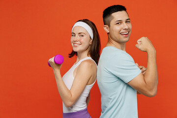Side view young fun fitness trainer instructor sporty two man woman in headband t-shirt hold dumbbells show muscles spend weekend in home gym isolated on plain orange background. Workout sport concept