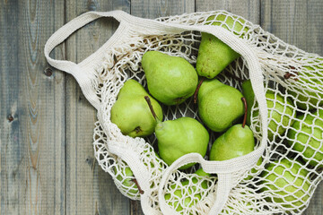 Indoors flat lay top view of a wooden rustic table on which lies a beige reusable shopping bag containing organic wholesome ripe green pears, space for text.