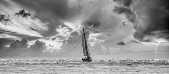 Rolgordijnen Black and white view of a sailboat navigating a silvery sea in a stormy cloudy sunset © Miguel Ángel RM