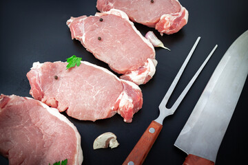 Pieces of raw roast beef meat for grilling on dark background with ingredients for cooking, knife and meat fork.