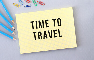 TIME TO TRAVEL text in notebook on gray background next to pencils and paper clips.