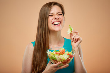 Happy laughing lady with closed eyes in azure dress eating green salad from glass bowl, isolated portrait on peach color background