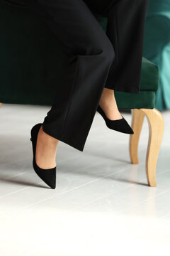 Cropped photo of legs in a classic black high heels shoes and black pants of a business woman sitting on a green chair by the window in the office.