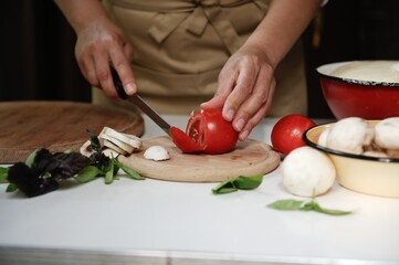 Chef's hands cut tomatoes on a chopping board with stacked sliced champignons, culinary aromatic herbs on kitchen table