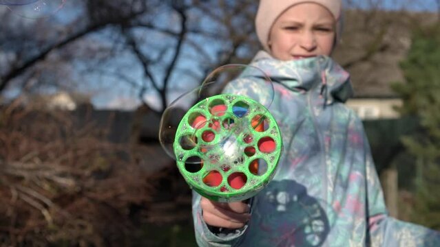 Slow motion of child girl in blue colorful jacket blowing soap bubbles toy gun