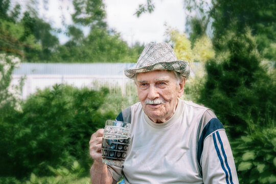 large portrait of a gray-haired old man in a hat with a glass of dark drink. man with a glass mug is sitting in the garden.