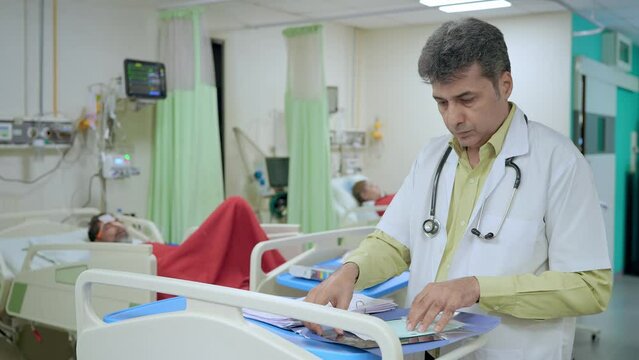 Middle Aged Asian Indian Male Physician Or Doctor Wearing  White Apron And Stethoscope Is Standing In An Intensive Care Unit Busy Examining Medical Report With Patients In The Back Of A Hospital Ward.