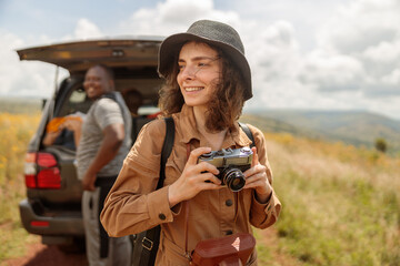 Smiling female traveler with photo camera enjoying the journey through the savannah next to safari car - obrazy, fototapety, plakaty