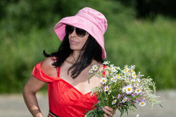 A girl in a pink hat, glasses in a red dress holds a bouquet of wild flowers in her hands on a blurred background. Close-up. Summer outdoor recreation.