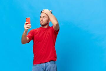 Self loved tanned handsome man in red t-shirt doing selfie video call using phone touch hair posing isolated on blue studio background. Copy space Banner Mockup. Online People lifestyle concept