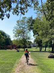 Boy rides his bike down a gravel path through a park wearing a backpack and helmet.