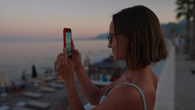 Portrait of woman using modern smartphone for recording video of amazing summer sunset over sea. Focus on mobile screen. Dark atmosphere around. High quality 4k footage