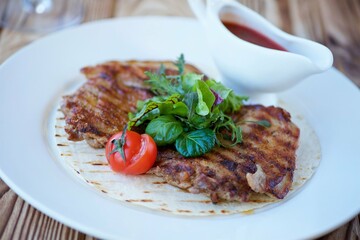 A piece of fried meat lies on a white plate. Decorated with vegetables and herbs. There is a red sauce nearby. The concept of recipes from the restaurant