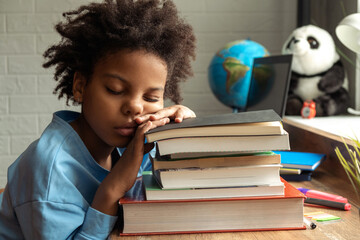 Tired African-American girl with closed eyes doing homework at home,puting her head on a stack of...