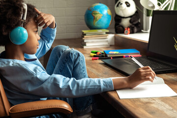 African-American girl doing homework,making notes,using headphones and laptop at home.Back to school concept.School distance education at home,home schooling,e-learning,diverse people.