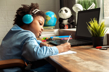 African-American girl doing homework,making notes,using headphones and laptop at home.Back to school concept.School distance education at home,home schooling,e-learning,diverse people.Copy space.