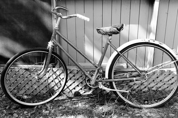 A black and white image of an old vintage bike leaning against a fence. 