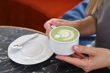 Fototapeta na wymiar Woman with a cup of matcha latte sits in the cafe. Delicious green matcha tea in a green cup close-up. Japanese hot milky beverage. Healthy Matcha Tea Made with Organic Ingredients