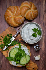 Spicy tzatziki with cucumber slices on fresh bun and chopped herb ingredients on cutting board.