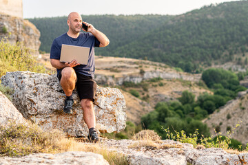Freelance man or digital nomad using a laptop and talking on the phone sitting on the rock of a mountain