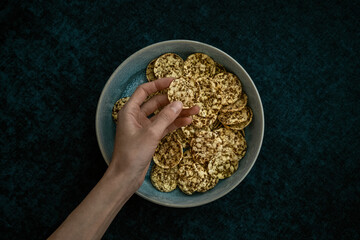 Woman's hand reaching out for round multigrain corn tortillas (crackers or chips) on a plate. Dark green background. Organic eating, detox diet. Healthy food snack concept.