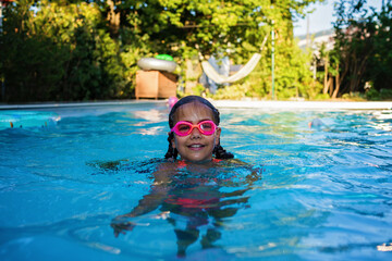 Beautiful girl with two braids in swimming suit and goggles smiles and enjoy training swim at pool in summer camp. Refreshing at heat weather, active vacation and healthy lifestyle, happy summertime