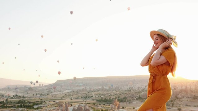 Young beautiful woman in elegant long dress in front of Cappadocia landscape in the sun with hot air balloons in the air. Turkey.