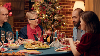 Senior couple talking to married young adults while enjoying traditional home cooked food at Christmas dinner. Festive people celebrating winter holiday with authentic meal and champagne.