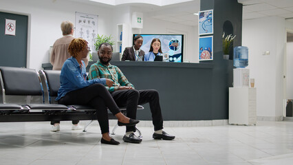 African american man and woman talking about disease in hospital reception lobby, waiting to attend consultation appointment with specialist. Patients in waiting area. Tripod shot.