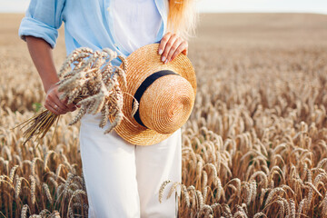 Close up of summer female accessory and clothes. Woman holding straw hat wearing linen shirt in wheat field