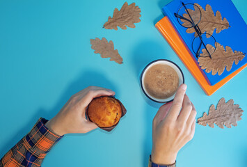 Woman's hand with cup of coffee, books and cake on a autumn blue background