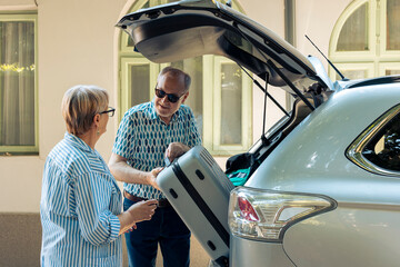 Elderly couple loading trolley in car trunk, travelling on retirement holiday vacation during...