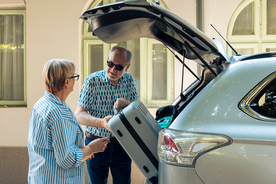 Elderly couple loading trolley in car trunk, travelling on retirement holiday vacation during summer. Senior people leaving on road trip adventure, putting suitcase and travel bags.