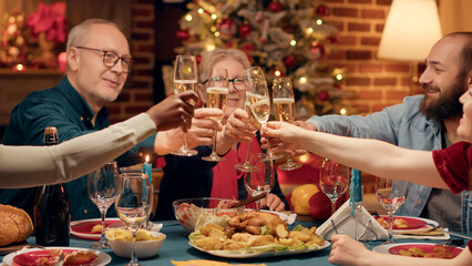 Festive diverse family members enjoying Christmas dinner at home while drinking champagne. Joyful multicultural people celebrating winter feast with home cooked food and sparkling wine.