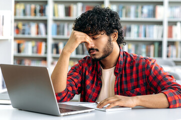 Exhausted sad indian or arabian male university student, in a stylish shirt, sits at a table in the library, closed his eyes, massages nose bridge, tired of boring studies, has a migraine, needs break