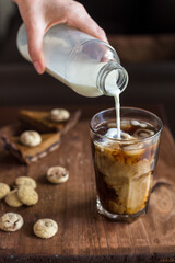 A woman's hand holds a bottle of milk and pours it into a glass of cold coffee. on the table in the background lies chocolate and biscuit cookies