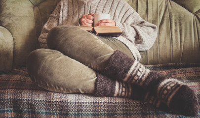 Woman sitting on sofa dressed in warm clothes holds a book and mug with hot drink. Hygge concept