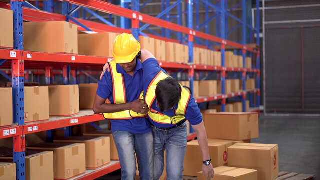 Careless Asian Warehouse Worker Lifting A Heavy Cardboard Box To The Shelf Then Some Of The Box Falling And Hit By Accident And His Friend Run To Safe Or Help Him. Safety Instruction In Working Area.