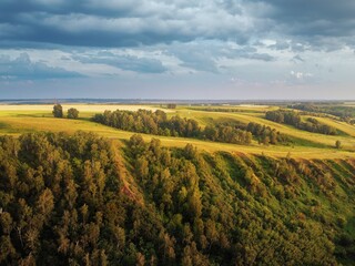 High hills with fields of grass and groves of birches. Erosion of clay hills. The light of the evening sun illuminates the clouds and the grass and trees
