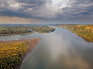 Riverbed under the mountain and groves of trees on the shore. Evening light of the sun. Clouds in the sky and reflection in the water. River Ob