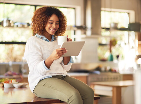 Relaxing done anywhere with the freedom of wireless technology. Shot of a happy young woman using a digital tablet and drinking coffee in the kitchen at home.