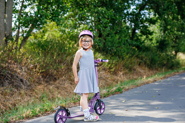 Fototapeta premium Portrait of active little preschool girl with glasses and helmet riding scooter on road in park outdoors on summer day. Seasonal child activity sport. Healthy childhood lifestyle